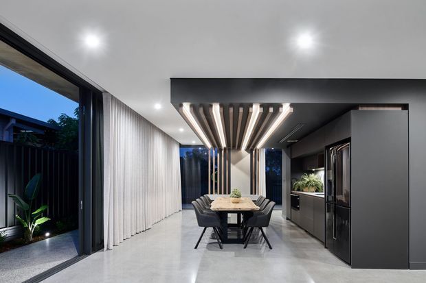 A refined contemporary open-plan dining and kitchen interior with polished light-grey flooring, dark built-in cabinetry, a large dining table, and black upholstered dining chairs. Full-height glazing with sheer curtains opens toward a planted courtyard, while a dramatic recessed ceiling feature combines dark panels with linear timber slats and integrated lighting.