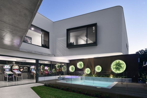 Contemporary two-storey residence with crisp white rendered walls, dark-framed windows and louvres, and a dramatic cantilevered concrete overhang. The courtyard-facing ground floor opens to a games or entertainment room with arcade machines, while a glass-fenced illuminated swimming pool and landscaped garden sit in the foreground. A dark feature wall with circular illuminated planting displays adds a strong architectural accent.
