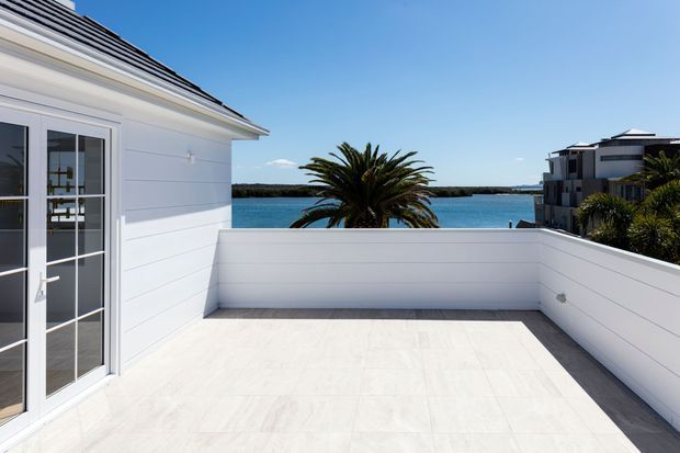 Bright rooftop or upper-level terrace overlooking a calm body of water. The terrace has pale tiled flooring, white horizontal wall cladding and white parapet walls, with a glazed French door opening into the building. A palm tree and neighbouring modern residences are visible beyond the terrace, creating a clean coastal setting.