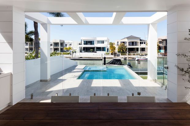 Bright contemporary dining area opening through floor-to-ceiling glazing onto a private pool terrace, with a canal-side residential setting beyond. The room features a large dark timber dining table, white structural framing, pale tiled flooring, and a strong indoor-outdoor connection.