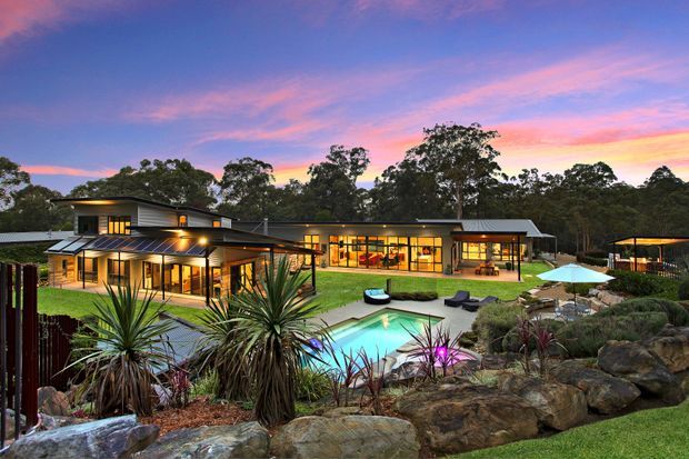 A wide exterior view of a low-profile contemporary residence at dusk, set within a landscaped bush setting. The home features broad glazing, multiple shallow roof planes, warm interior lighting, covered outdoor living areas, a swimming pool, lawn, rock landscaping, and sculptural drought-tolerant planting.
