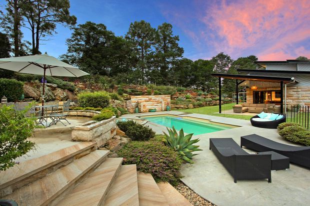A landscaped residential outdoor entertaining area with a turquoise swimming pool, broad paved terrace, stepped stone garden beds, outdoor dining under a large umbrella, and black lounge furniture. A low modern pavilion with stone-clad walls and a dark roof sits beside the pool, surrounded by mature trees and layered planting.