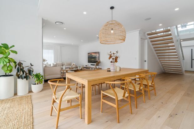 Bright, open-plan dining area with a large light timber dining table and matching woven-seat chairs, opening onto a relaxed living room. The space features pale timber flooring, white walls and ceiling, abundant recessed lighting, indoor plants, a woven rattan pendant light, and a timber-and-white open staircase with a skylight above.