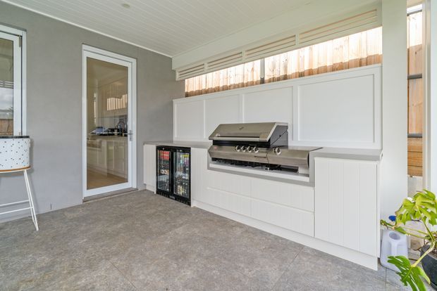 A bright, enclosed outdoor entertaining area with a built-in stainless-steel barbecue, white panelled cabinetry, under-counter beverage refrigerators, and a long horizontal clerestory window. The space has grey tiled flooring, light grey walls, and a white-lined ceiling, with a glazed door connecting to the interior.