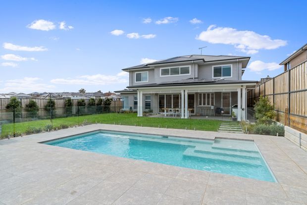 A two-storey suburban home with a grey weatherboard-style exterior, low-pitched hipped roof and covered rear patio. The property opens onto a broad paved terrace with a rectangular swimming pool, manicured lawn and timber boundary fencing.