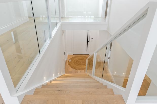 Bright contemporary stairway viewed from above, with pale timber stairs, white walls and structural trim, clear glass balustrades, and abundant natural light from large windows and a double-height opening. A lower-level hallway with a round rug is visible below.