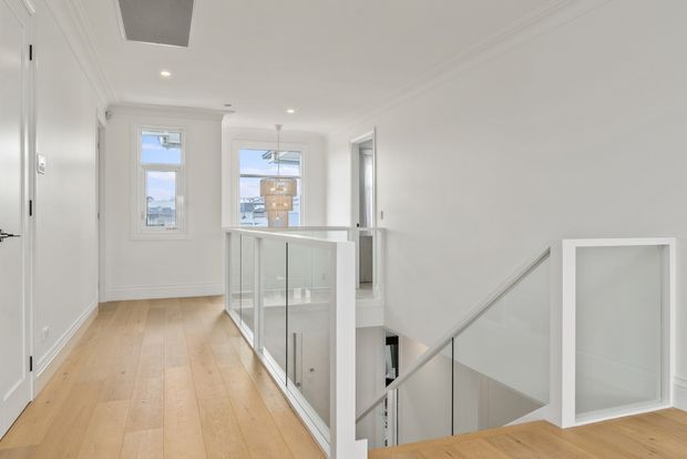 Bright, minimalist upper-level landing or hallway overlooking a stairwell. The space features white walls and ceiling with simple cornice detailing, pale timber flooring, a frameless glass balustrade with slim white supports, recessed downlights, and a white-framed opening or door at the stair landing. Tall windows provide daylight and a view of the surrounding urban setting.