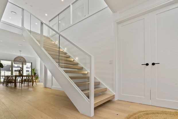 Bright contemporary residential interior showing an open stairway with light timber treads, white painted balustrade and glass panels. White paneled double doors and built-in cabinetry line the right wall, while a dining area with timber flooring and pendant lighting is visible beyond the stair. The space has a clean, airy, refined appearance with strong white and pale wood finishes.