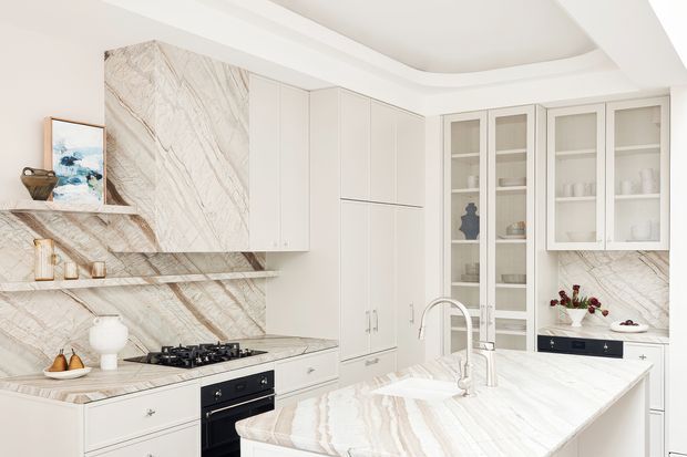 Bright, refined kitchen featuring extensive white cabinetry, a large pale veined stone island and matching splashback, a black gas cooktop and oven, glass-front display cabinets, and a decorative recessed ceiling detail. The space has a clean, luxurious appearance with warm neutral accents and integrated storage.