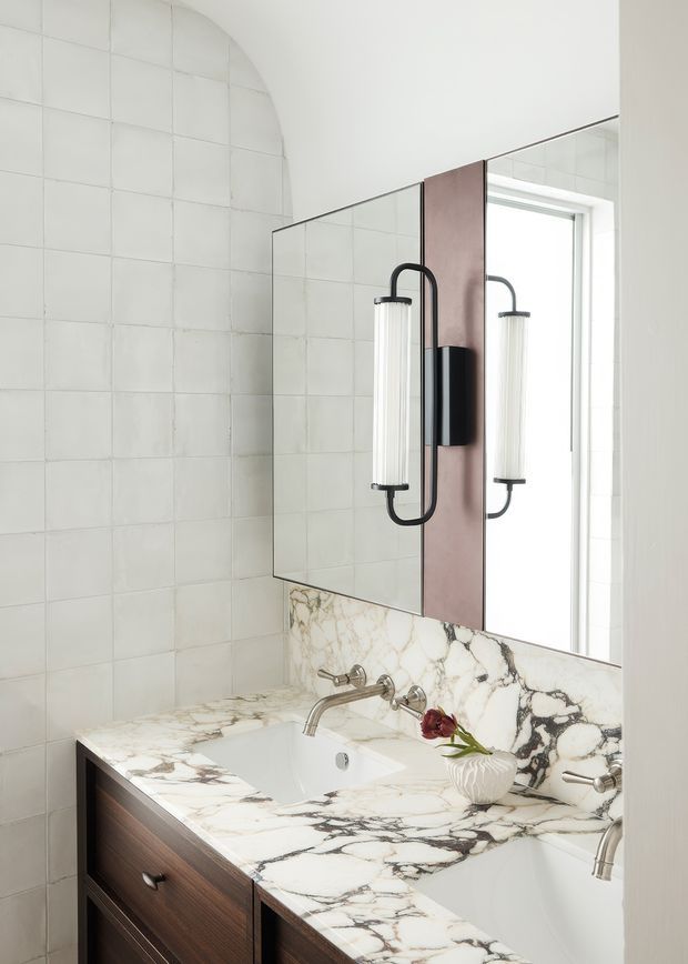 A bright, refined bathroom vanity with a double sink, white marble countertop and backsplash featuring bold grey and brown veining, dark timber cabinetry, and a large mirrored wall. Sculptural black-and-white wall sconces flank a central timber-toned mirror panel, while pale tiled walls and a window create an airy atmosphere.
