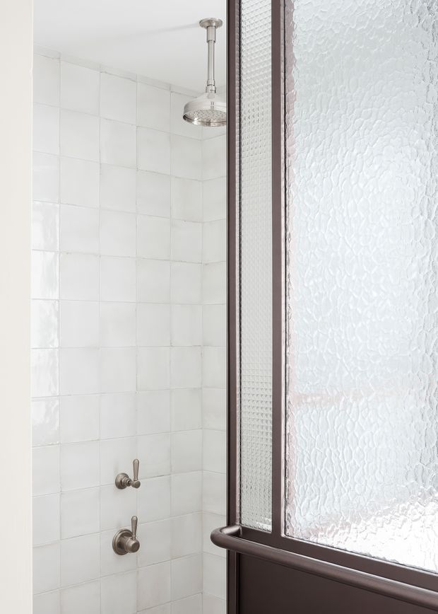 A close-up view of a compact shower enclosure. The shower walls are finished with glossy white square tiles, with a chrome rainfall showerhead and matching controls. A dark brown-framed frosted textured-glass door occupies the right side of the image, allowing diffused light into the space.