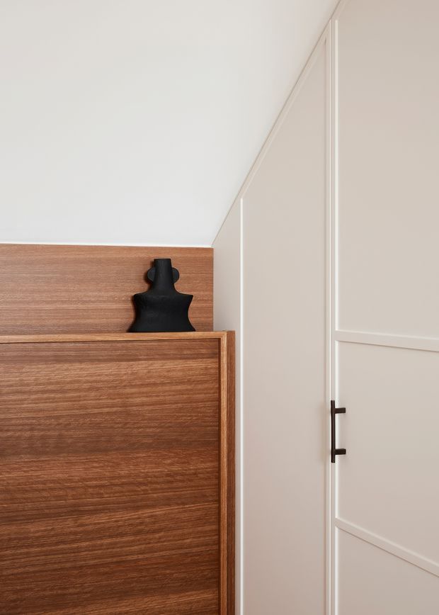 Close-up interior detail of a refined built-in timber cabinet or sideboard beside a tall cream-painted panelled door. The cabinet has a warm medium-to-dark wood veneer with horizontal grain, topped by a matching timber ledge displaying a small matte-black sculptural object. The adjacent door features recessed rectangular panels, slim trim, and a dark metal vertical handle. The composition is minimalist and warm, with clean contemporary joinery and soft neutral tones.