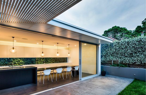 Contemporary covered outdoor entertaining area with a fully open glazed kitchen and dining space. A long island bench, linear fixed cabinetry, pendant lights and bar seating sit beneath a slatted pergola and broad roof overhang. The space opens onto a paved courtyard with raised garden beds and screening vegetation.