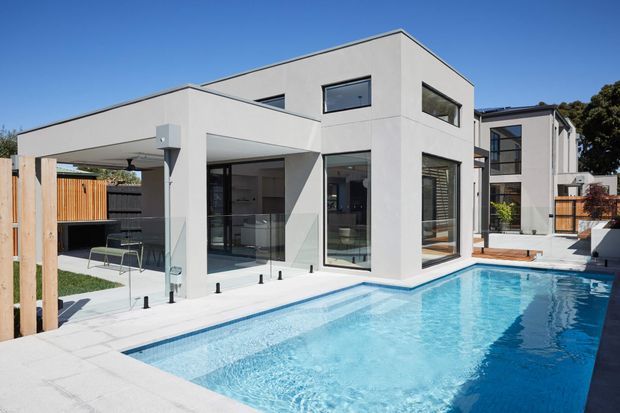 Modern two-storey residential home viewed from the pool area, featuring crisp white rendered cubic volumes, extensive dark-framed glazing, a covered outdoor entertaining area with glass balustrades, timber screening, pale paving, and a bright blue swimming pool.