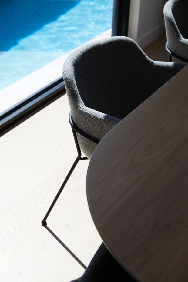 Close-up interior view of a round timber dining table with dark grey upholstered dining chairs beside full-height glazing. A bright blue swimming pool is visible outside, and strong sunlight falls across the pale floor.
