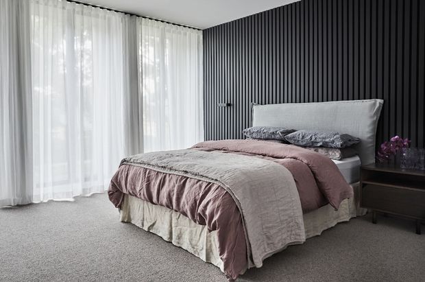 A contemporary bedroom with a neatly made bed layered in muted blush, taupe, and grey bedding. Floor-to-ceiling sheer curtains provide soft natural light, while a dramatic black vertical slatted feature wall forms the backdrop to the upholstered headboard. The room includes grey carpeting and a dark bedside table with flowers and decorative objects.