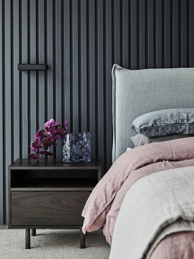 Close-up interior photograph of a sophisticated bedroom corner, featuring a bed with layered grey, blush pink and white bedding beside a dark timber bedside table. The backdrop is a dramatic charcoal vertical fluted wall panel, with a purple orchid and textured glass vessel providing decorative accents. The palette is moody, refined and contemporary.