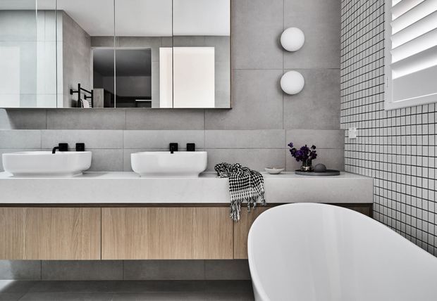 Contemporary bathroom with a double floating vanity, white vessel basins, timber-look cabinetry, large grey wall tiles, a small-format tiled feature wall, mirrored cabinetry, wall sconces, and a freestanding white bathtub. A shuttered window provides natural light, while restrained styling gives the room a clean, refined feel.