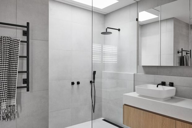 Contemporary minimalist bathroom with large light-grey wall and floor tiles, a frameless glass shower enclosure, matte black shower fittings and towel rail, floating timber-veneer vanity, white vessel basin, mirrored cabinetry, and a skylight.