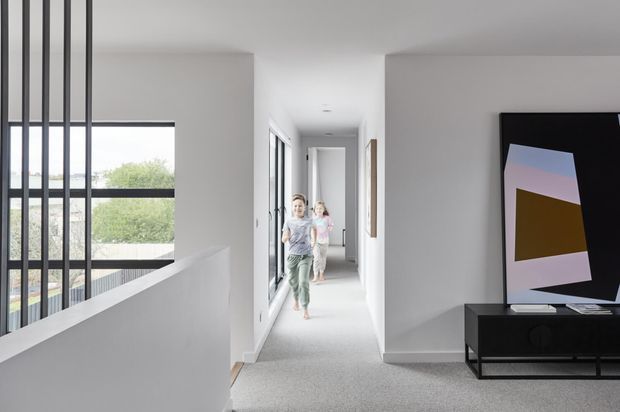 Bright contemporary interior hallway with a carpeted floor, white walls and ceiling, full-height glazing along the left side, a half-height stair balustrade, and slim black vertical screen elements. Two children are visible in the corridor, with a black media console and abstract artwork in the foreground.