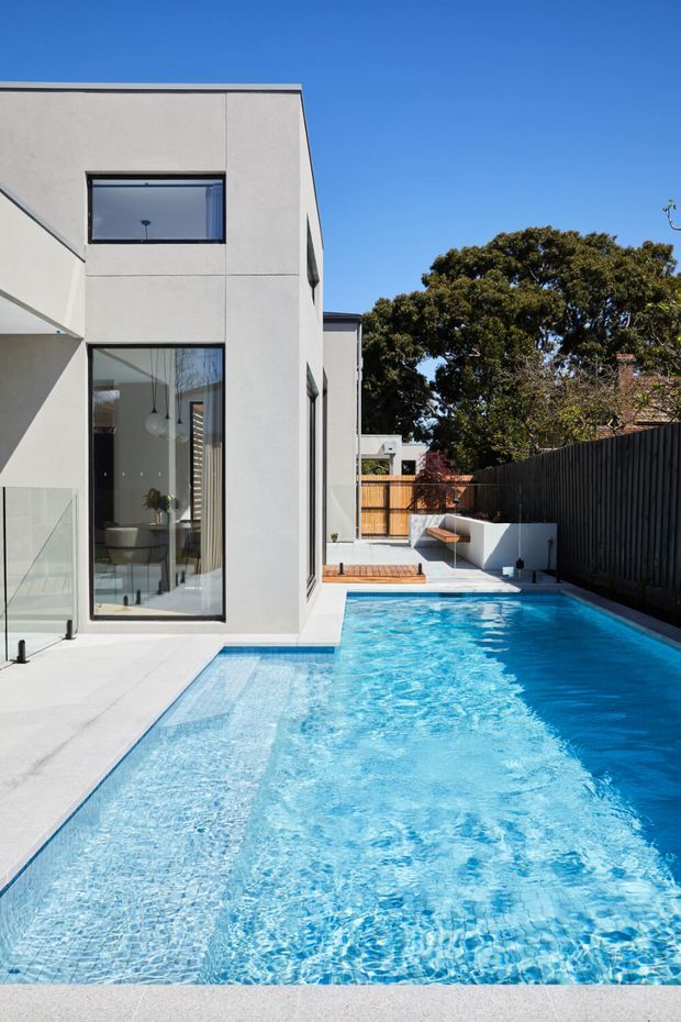 Exterior architectural photograph of a contemporary multi-level home with a clean white rendered facade, large dark-framed windows, and a long narrow swimming pool running alongside the building. The pool is bordered by pale paving, a dark vertical fence, built-in white seating, and timber screening. Bright clear daylight and strong geometric lines emphasize the modern design.