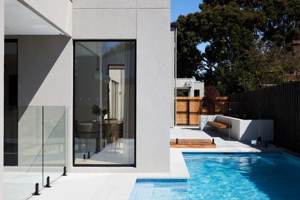 Contemporary residential exterior showing a crisp white rendered multi-level facade with large dark-framed glazing, a glass balustrade, and a bright blue swimming pool beside a timber deck and built-in outdoor seating. Mature trees and a dark privacy fence surround the compact outdoor area.