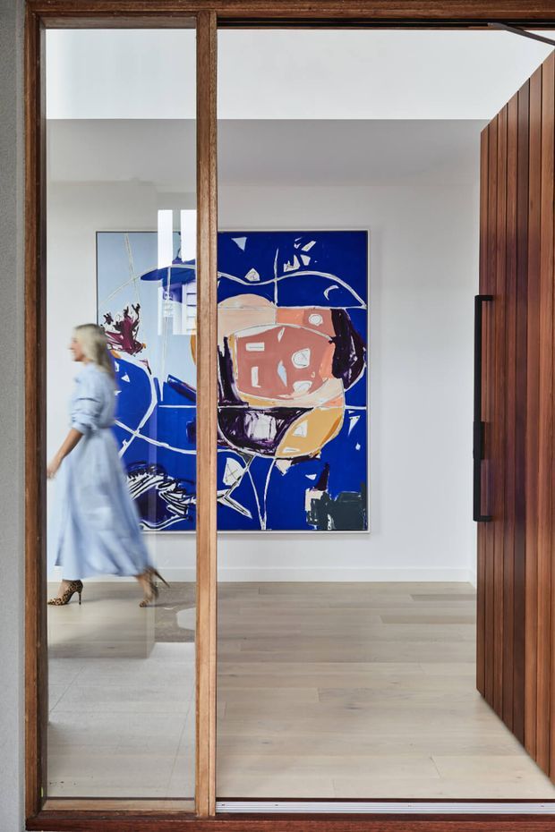 A refined interior entry or hallway viewed through a large timber-framed glazed doorway. The space has pale wide-plank timber flooring, white walls and ceiling, a sculptural vertical timber door or wall panel on the right, and two large bold abstract artworks in blue, white, coral and black. A person is walking through the left side of the scene.