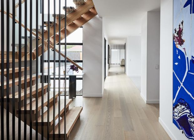 Bright contemporary hallway and stairway interior with pale timber flooring, a sculptural open-riser timber staircase, and black vertical metal balustrades. White walls and abundant glazing create an airy feel, while a large colourful artwork provides a bold accent.