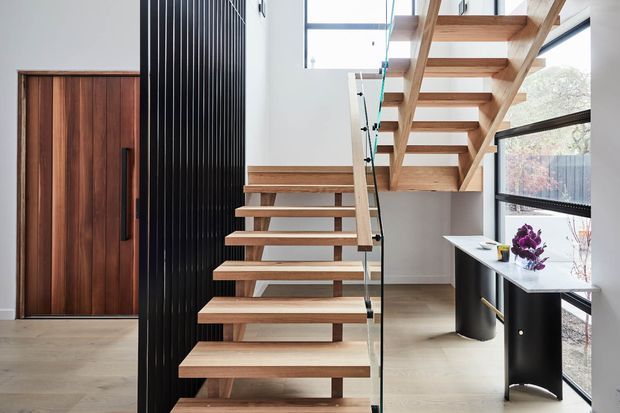 Bright contemporary interior featuring an open-riser timber staircase with glass balustrade, a tall black vertical-panel feature wall, a dark timber entry door, and a compact black-and-white built-in workspace beside expansive glazing.