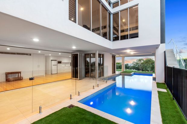 A bright contemporary multi-level home centered around a narrow swimming pool courtyard. The image shows white rendered walls, extensive floor-to-ceiling glazing, upper-level balconies, warm timber flooring and ceilings, a compact kitchen visible through the glass, an external stair, and manicured artificial grass around the pool.