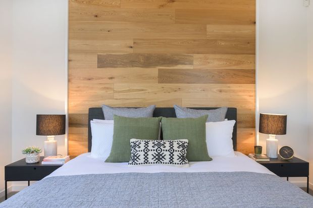 A styled bedroom with a large bed centered against a full-height natural timber feature wall. The bed has white bedding, muted sage-green and grey pillows, and a patterned accent cushion, flanked by black bedside tables with lamps and small decor. The overall look is warm, contemporary, and restrained.