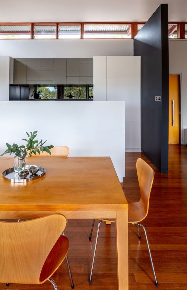 A bright, modern open-plan dining area with a large light timber dining table and matching sculptural chairs in the foreground. Behind it is a minimalist white kitchen with streamlined handleless cabinetry, a dark benchtop or lower cabinet zone, and a white dividing wall. Warm timber flooring, high clerestory windows with timber frames, and a dramatic dark charcoal partition wall frame the space, with a vivid orange doorway adding contrast.