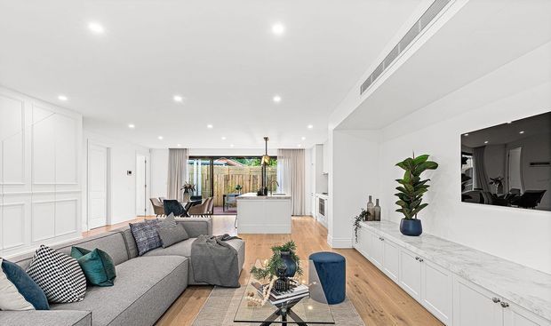 Bright open-plan living room with a large grey sectional sofa, patterned cushions, timber flooring, white paneled built-in cabinetry, a marble-look entertainment and storage wall, and a compact white kitchen island in the background. Recessed ceiling lighting, sheer curtains, indoor plants, and contemporary furnishings create a refined, airy interior.