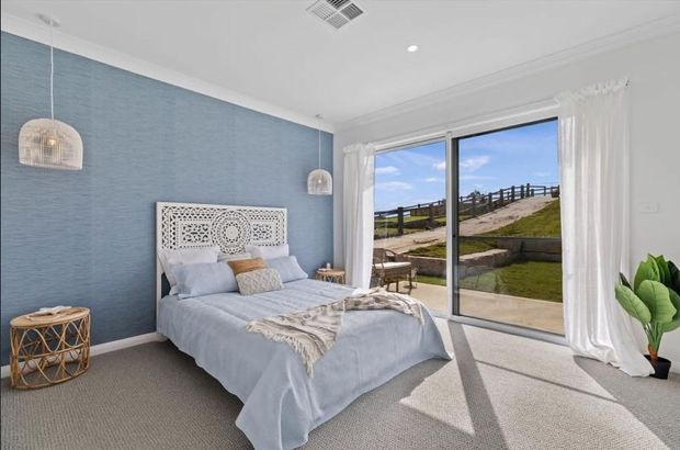 Bright coastal-style bedroom with a pale blue textured feature wall, white decorative headboard, layered light-blue bedding, woven pendant lights, and soft white curtains framing full-height sliding glass doors. The doors open to a landscaped outdoor area with a walkway and timber fence, while a neutral carpet and white ceiling create an airy, relaxed interior.