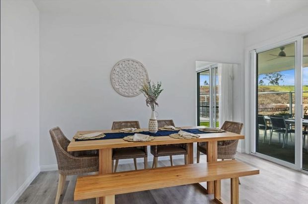 Bright, airy dining room with a long solid timber dining table and matching bench, woven dining chairs, white walls, light timber-look flooring, and large sliding glass doors opening to an outdoor entertaining area. A simple round wall artwork and vase provide understated decoration.