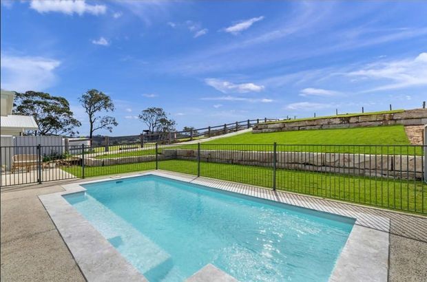A bright outdoor swimming pool in a landscaped residential setting, enclosed by a black metal fence. The pool has pale blue water and a light grey paved surround, with a manicured sloping lawn, stone retaining wall and timber fencing beyond. A small portion of a light-coloured building is visible at the edge of the frame under a vivid blue sky.