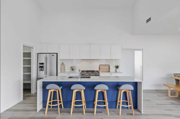 Bright contemporary open-plan kitchen with white flat-panel cabinetry, a large island featuring a vivid blue front, and four timber-and-white bar stools. Stainless steel appliances, pale timber-look flooring, and minimal white walls create a clean, airy setting. A dining area is partially visible to the right and a pantry is visible through the opening on the left.