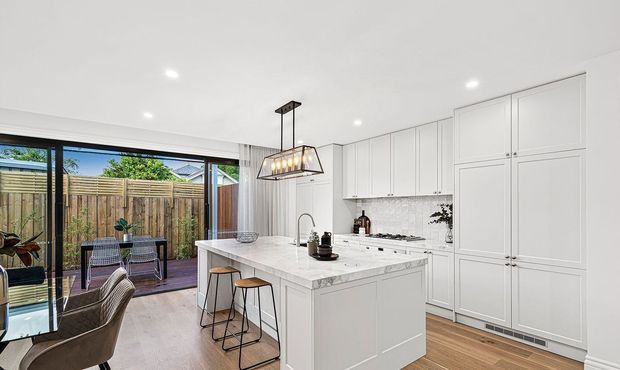 Bright, contemporary white kitchen with a large central island, integrated tall cabinetry, marble-look benchtops, tiled splashback, timber flooring, and a linear pendant light. The kitchen opens through large sliding glass doors to a small outdoor deck and fenced courtyard.