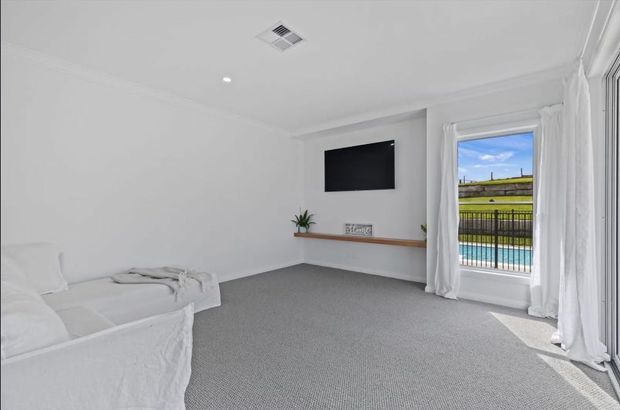 Bright, minimalist living room with white walls, white slipcovered seating, grey carpet, a wall-mounted television, and a simple timber display shelf. Full-height glazed doors with sheer curtains open to a balcony overlooking a swimming pool.