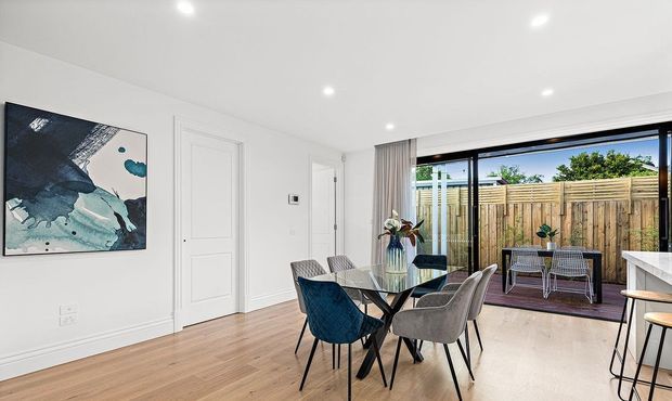 Bright contemporary open-plan dining area with a timber floor, white walls and ceiling, recessed downlights, and a large black-framed sliding glass opening to a timber-decked outdoor space. A glass dining table with upholstered chairs occupies the centre, while the edge of a kitchen island is visible at right. The room has clean-lined, neutral styling with a blue abstract artwork and sheer curtains.