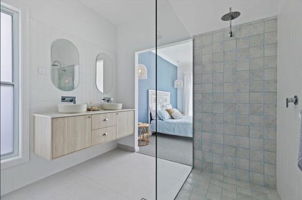 Bright contemporary bathroom with a floating light-wood vanity, white vessel basins, rounded mirrors, pale tiled walls and floor, and a glass-enclosed walk-in shower with blue-grey square tiles. A bedroom is visible through the open doorway in the background.