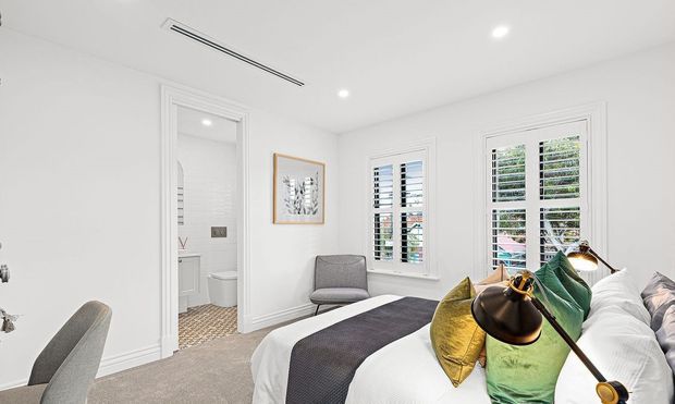 Bright, refined bedroom with white walls and traditional decorative trim, plantation-shuttered windows, a neatly made bed with layered white, grey, green and mustard bedding, a grey accent chair, framed artwork, and a small adjoining bathroom visible through the open doorway.