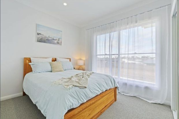 Bright, airy bedroom with a timber bed frame and pale blue bedding. The room has white walls, light carpet, sheer full-height curtains, a bedside lamp, and a large window overlooking an exterior setting. A soft landscape artwork provides a subtle decorative accent.