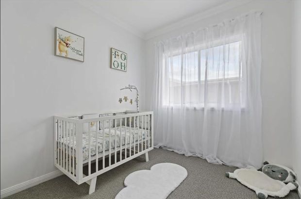 Bright nursery with a white timber cot, soft grey carpet, sheer white curtains over a large window, and gentle animal-themed decor. The room has a calm, neutral palette and traditional trim details.