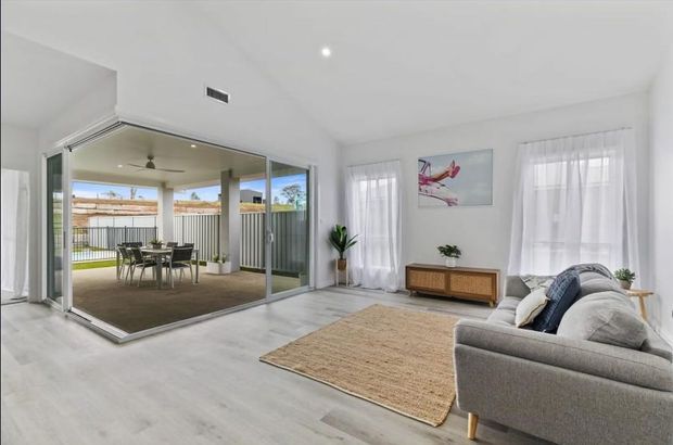 Bright contemporary living room with pale timber-look flooring, white walls and ceiling, a large grey sofa, woven neutral rug, timber sideboard, indoor plants and abstract artwork. Full-height sliding glass doors open to a covered outdoor dining area and fenced yard, creating a strong indoor-outdoor connection.