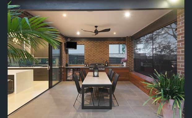 Enclosed indoor-outdoor dining and entertaining area with exposed red brick walls, large glazed openings, a tiled floor, timber cabinetry, and a long dining table with black chairs. A ceiling fan, recessed lighting, and abundant potted tropical foliage contribute to a contemporary industrial feel. A kitchen is visible through the glazing on the left.