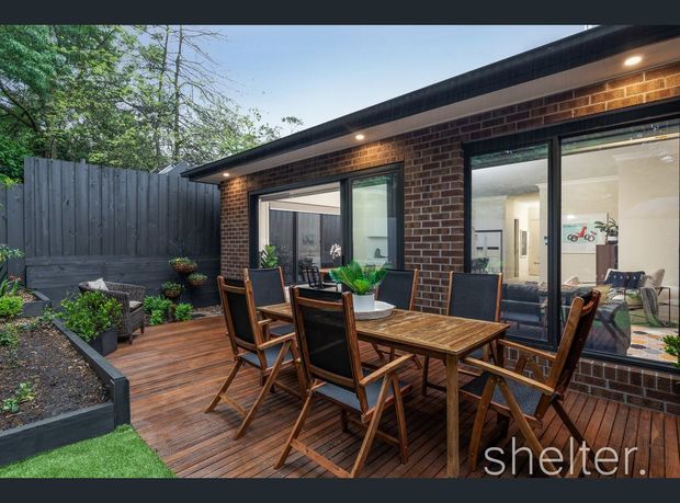 A compact landscaped courtyard and timber deck beside a single-storey brick extension. The deck features a large outdoor timber dining table with folding chairs, raised garden beds, potted plants, dark fencing, and large glazed openings looking into a bright interior. Recessed exterior lighting illuminates the brickwork and roof eaves.