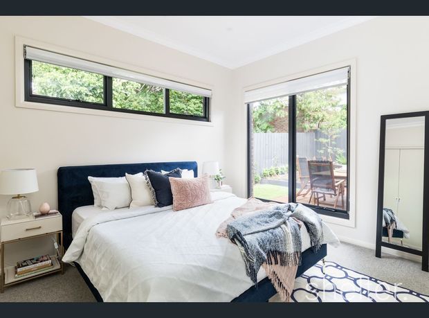 Bright contemporary bedroom with a navy upholstered bed, layered white and blue bedding, bedside tables, and a large black-framed floor mirror. Corner windows provide views to a small outdoor patio, while light neutral walls and carpet create an airy, comfortable atmosphere.