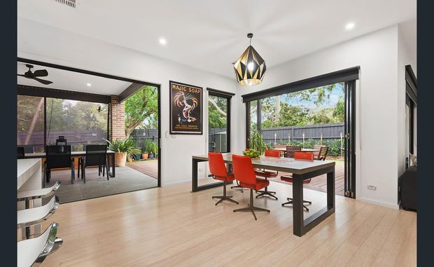 Bright contemporary dining area with a large timber dining table, red upholstered chairs, pendant lighting, pale timber flooring, and extensive black-framed glazing. An adjoining outdoor entertaining area and garden are visible through the open sliding doors, with exposed brick detailing and indoor plants adding warmth.