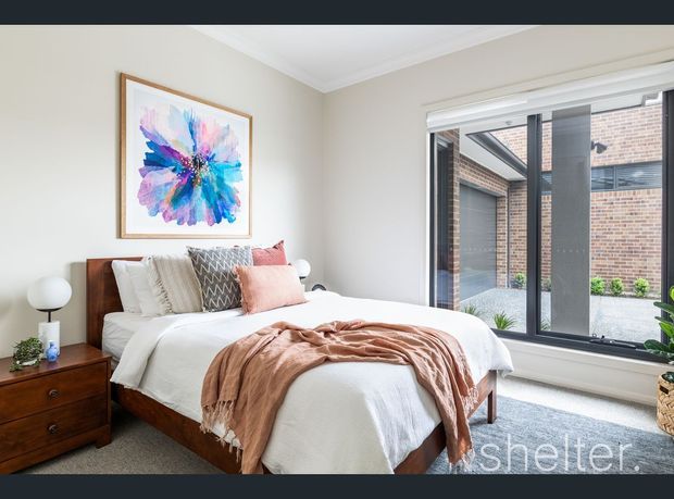 Bright, comfortably styled bedroom with a timber bed frame and matching bedside table, layered white bedding, blush and patterned cushions, and a soft terracotta throw. A colourful abstract floral artwork hangs above the bed. Large black-framed windows overlook a brick-lined courtyard, bringing in abundant natural light.